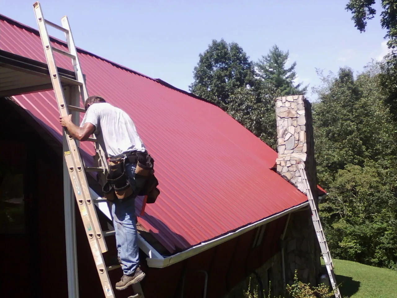 Roofer on a ladder beside a red metal roof and stone chimney — Soffit & Fascia Repair by Roofing Brothers in Sparr